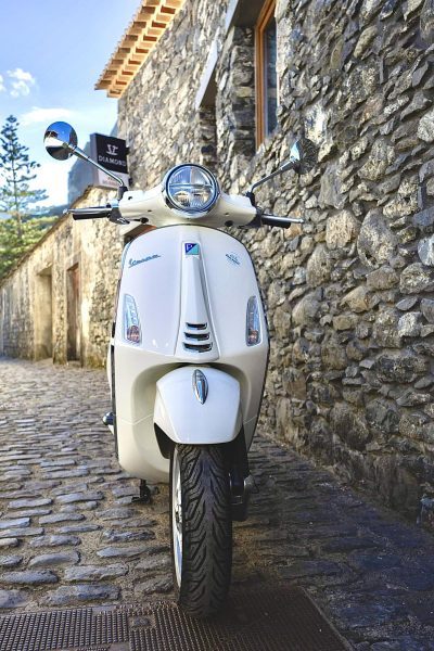 A white Vespa scooter parked near a stone house in madeira Island.The vehicle is a Vespa Primavera/GTS 125cc model, the type offered for rent to explore the island’s mountain roads and scenic viewpoints by Vespa Madeira Scooter Rent.