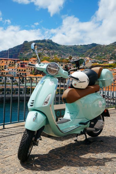 A green Vespa scooter is parked in a road and in the back Camara de Lobos. The vehicle is a Vespa Primavera/GTS 125cc model, the type offered for rent to explore the island’s mountain roads and scenic viewpoints by Vespa Madeira Scooter Rent.