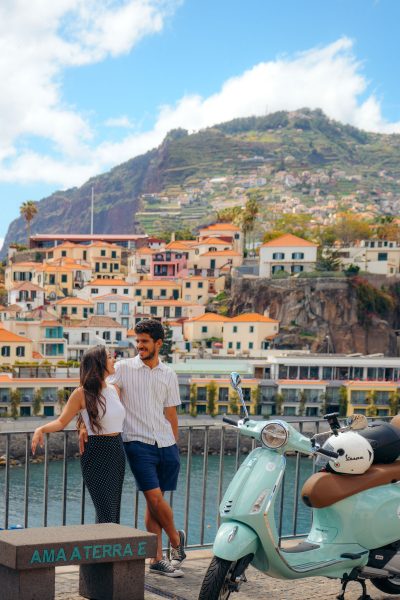 A couple hugging and smiling to eachother near a green Vespa that is parked, and in the background Camara de lobos. The type of scooter offered for rent to explore the Madeira island’s winding mountain roads and scenic viewpoints by Vespa Madeira Scooter Rent.