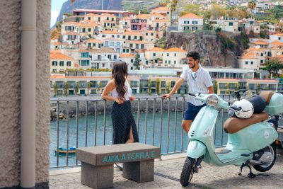 Vespa Scooter Guided City Tour at Câmara de Lobos where a couple is smiling to eachother near a green Vespa that is parked, and in the background Camara de lobos. The type of scooter offered for rent to explore the Madeira island’s winding mountain roads and scenic viewpoints by Vespa Madeira Scooter Rent.