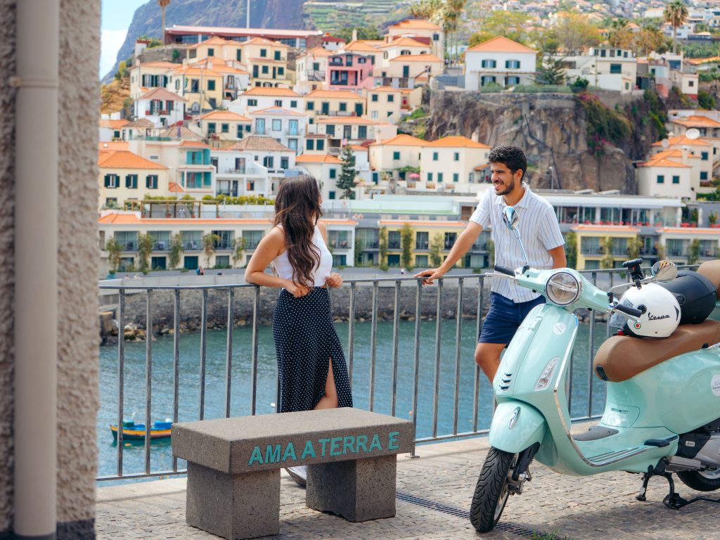 Vespa Scooter Guided City Tour at Câmara de Lobos where a couple is smiling to eachother near a green Vespa that is parked, and in the background Camara de lobos. The type of scooter offered for rent to explore the Madeira island’s winding mountain roads and scenic viewpoints by Vespa Madeira Scooter Rent.