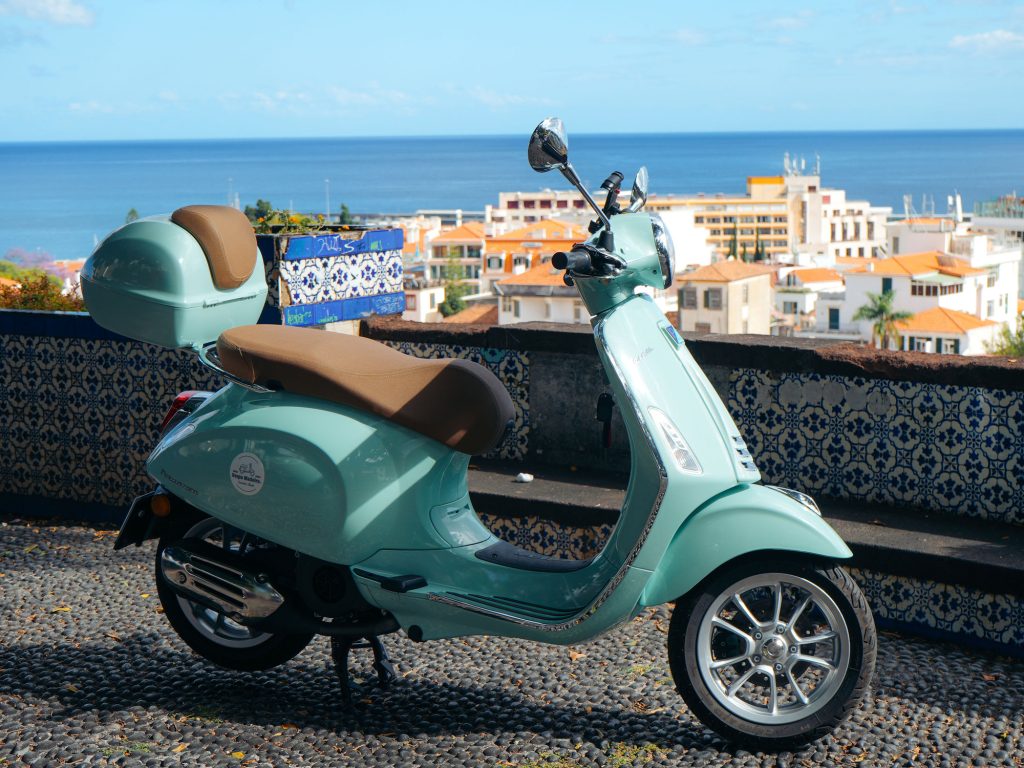 A green Vespa scooter is parked on a stone terrace, a funchal viewpoint in Madeira. The vehicle is a Vespa Primavera/GTS 125cc model, the type offered for rent to explore the island’s mountain roads and scenic viewpoints by Vespa Madeira Scooter Rent.