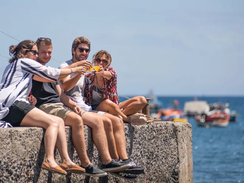 Friends toasting poncha drinks while relaxing by the sea during the Bar hopping and Poncha Workshop, an experience by Wine Tours Madeira.